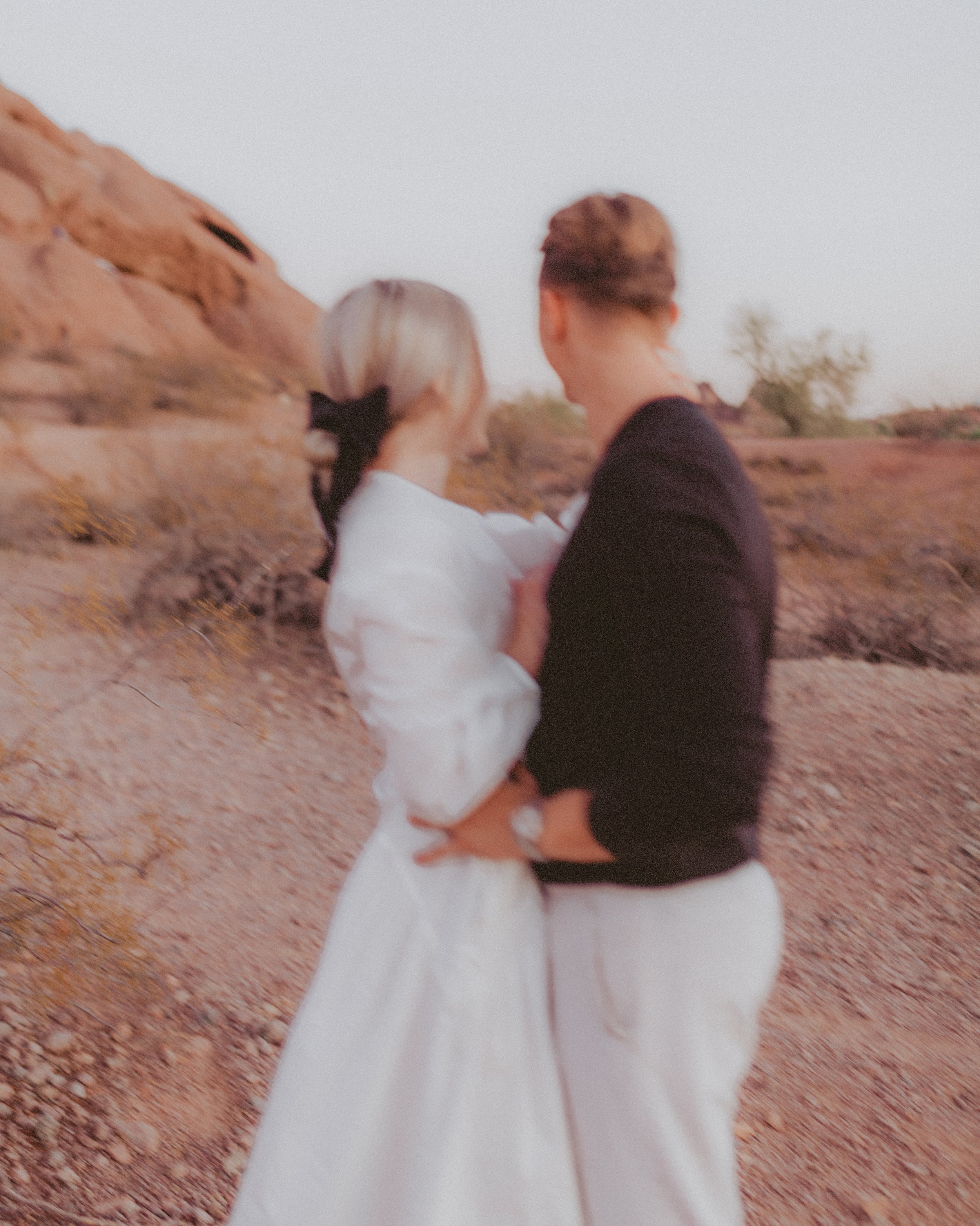 A blurry photo of a couple standing outdoors in a desert-like landscape, facing each other. The woman is wearing a white dress and a black hair bow, and the man is dressed in a black shirt and white pants. They are holding each other closely.