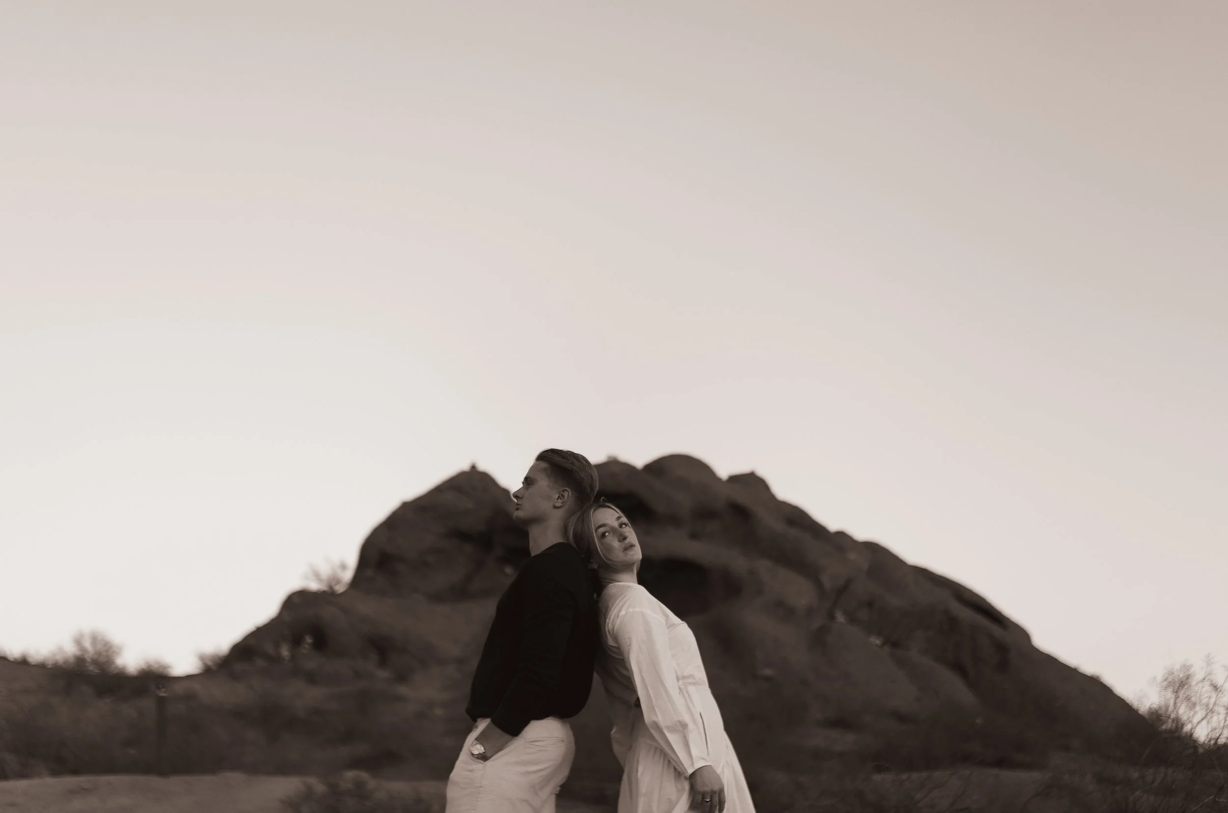 A black-and-white photograph of a man and woman leaning back-to-back in front of a rocky hill under a clear sky.