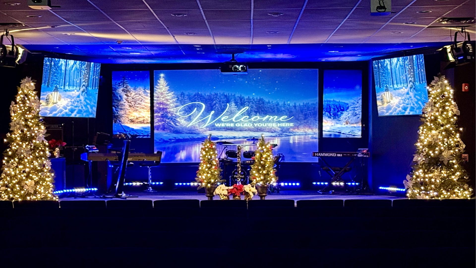 Stage decorated for Christmas with two lit Christmas trees, poinsettia flowers, and a large screen displaying a snowy winter scene and the words 'Welcome, We're Glad You're Here.' Musical instruments and a keyboard are on the stage.