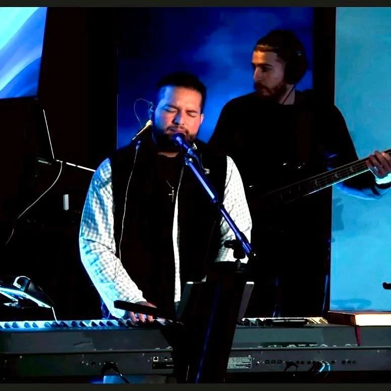 A male musician with eyes closed singing into a microphone while playing a keyboard, with another male musician playing a bass guitar behind him in a dimly lit setting.