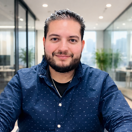 A man with dark hair, beard, and mustache smiling at the camera in a modern office space with glass walls and plants.