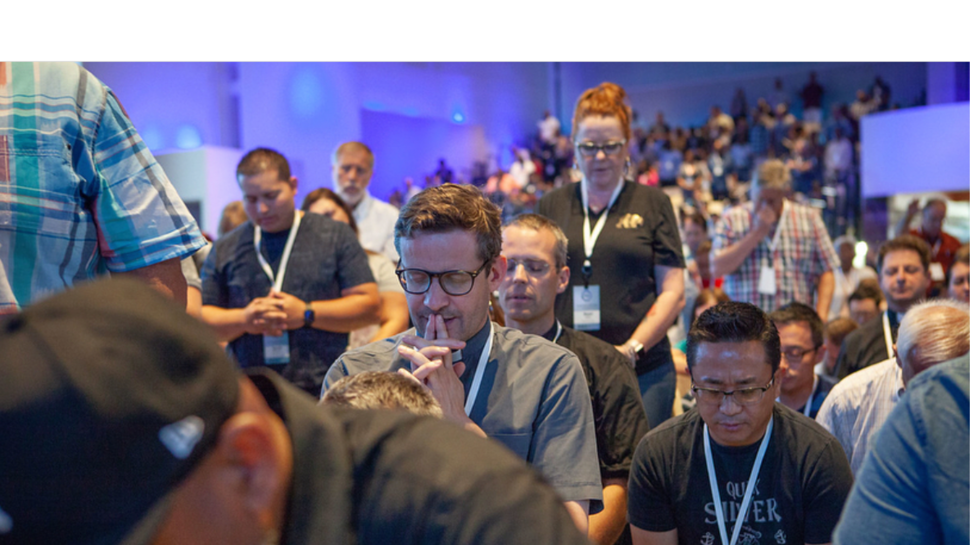 People praying and worshiping at a conference, with some standing and others kneeling, in a large indoor venue with blue lighting.
