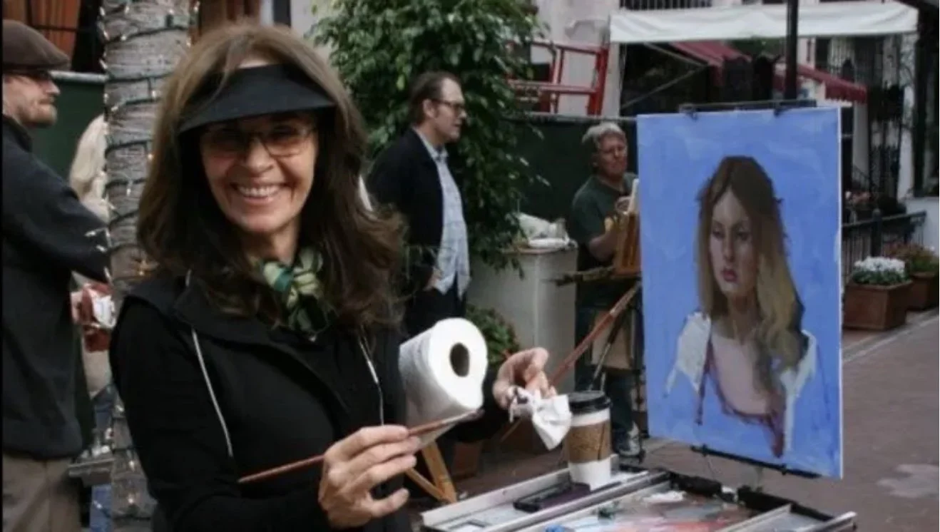 A woman smiles as she paints a portrait of a young woman on canvas outdoors. She is holding a paintbrush and a paper towel, with an easel, palette, and coffee cup nearby. Other people are also visible in the background, along with trees and buildings.