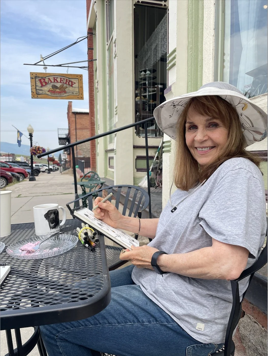 A woman with long reddish hair, wearing a wide-brimmed hat and gray T-shirt, sitting outside at a table, sketching in a notebook. On the table are pens, a mug with a dog image, and a glass plate with pink and white icing. Background shows a bakery sign and parked cars.