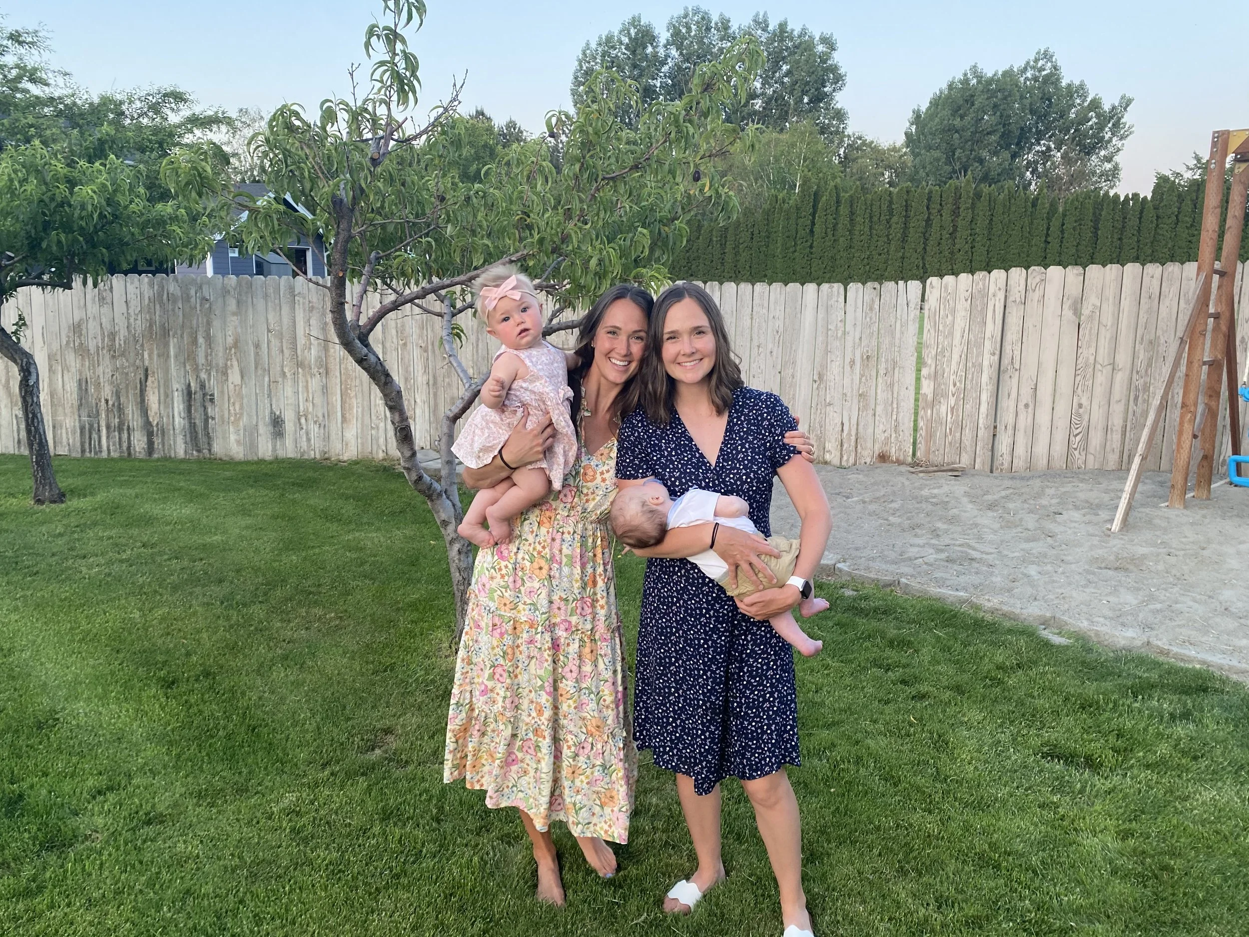 Two women standing on a green lawn holding two young children, one girl and one baby, smiling, with a wooden fence and trees in the background.