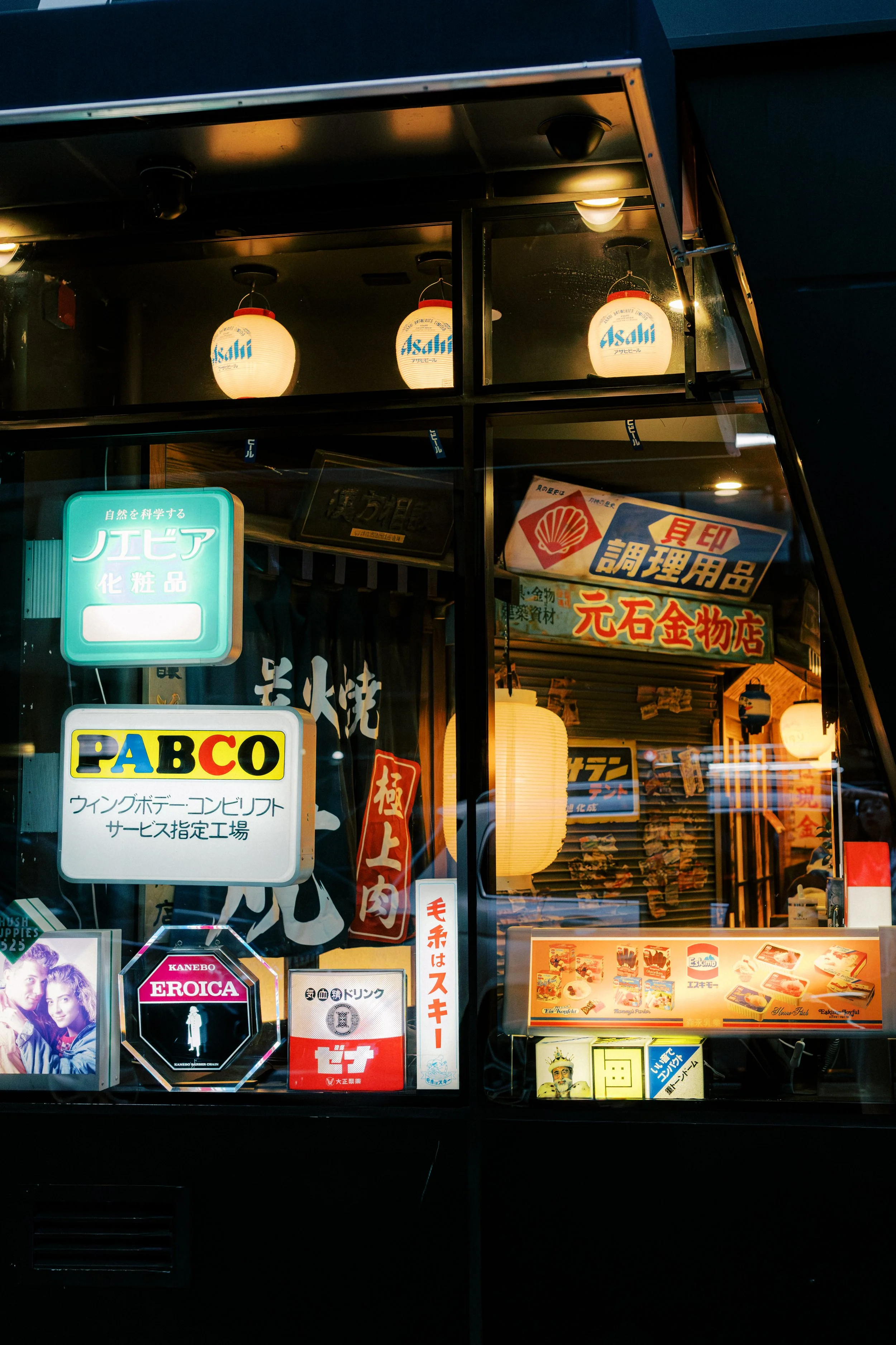 View of a storefront with Japanese signs, paper lanterns, and various advertisements in Japanese and English, illuminated at night.