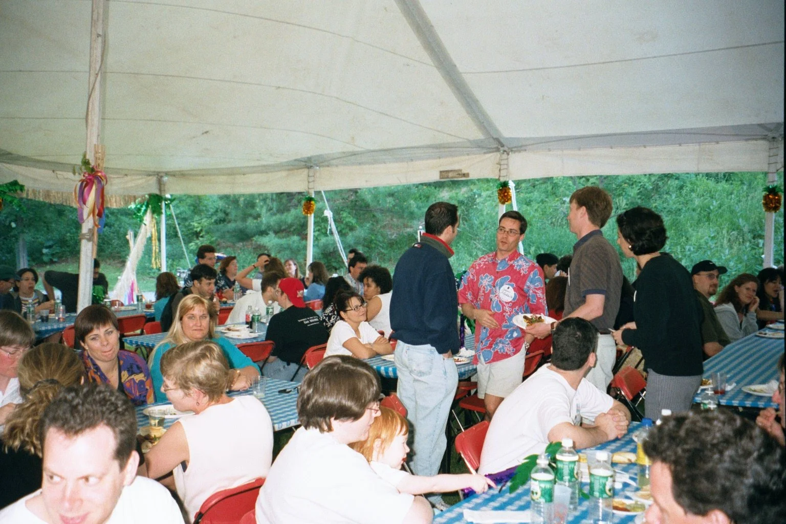 People gathered at a large outdoor event under a tent, sitting at tables with blue checkered tablecloths, and engaging in conversation.