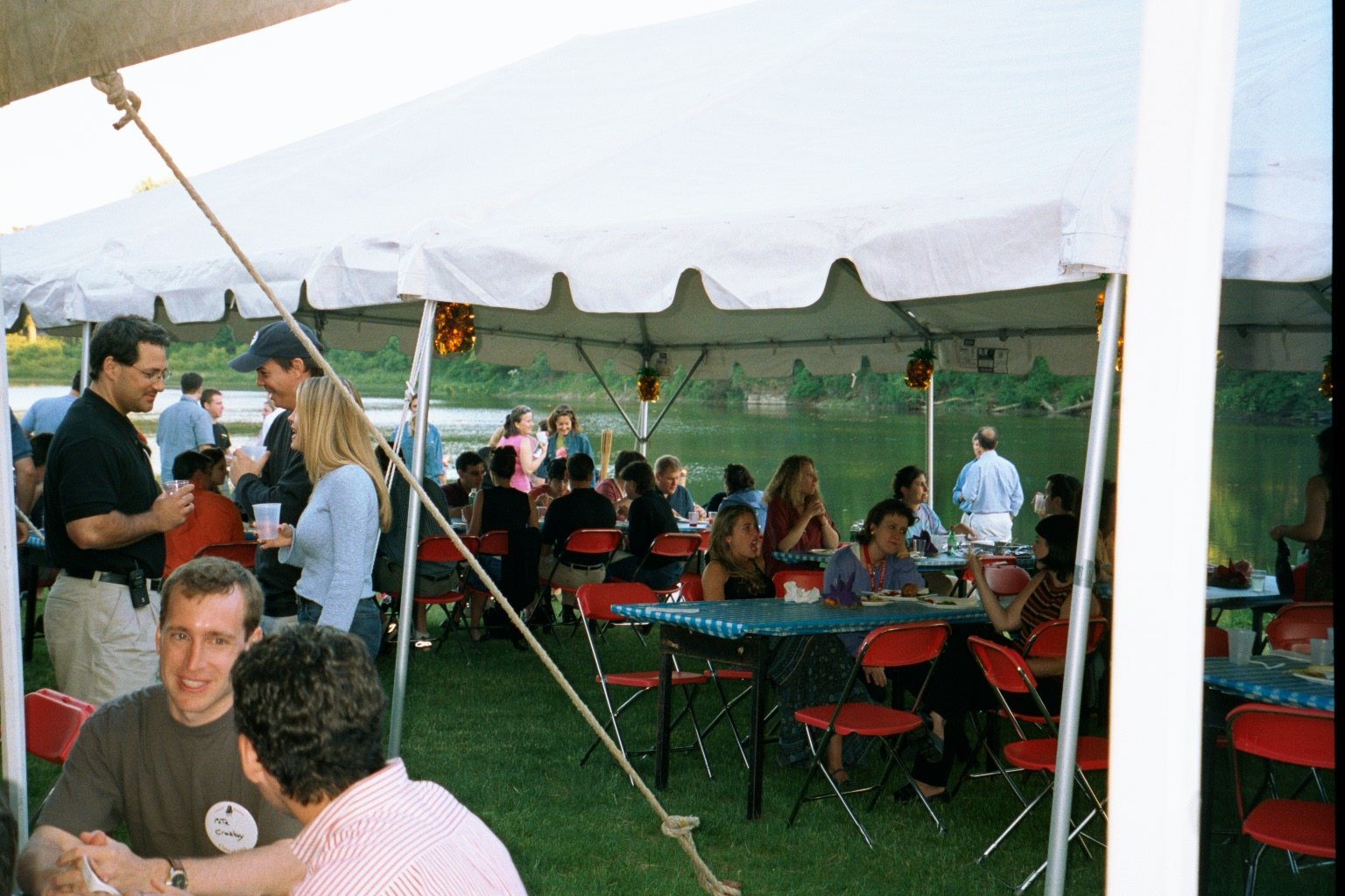 People gathered at an outdoor party under a white tent near a body of water, socializing and dining at tables with red chairs and blue checkered tablecloths.