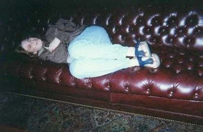 Child lying on a red leather tufted sofa, wearing a dark top, white pants, and blue shoes.