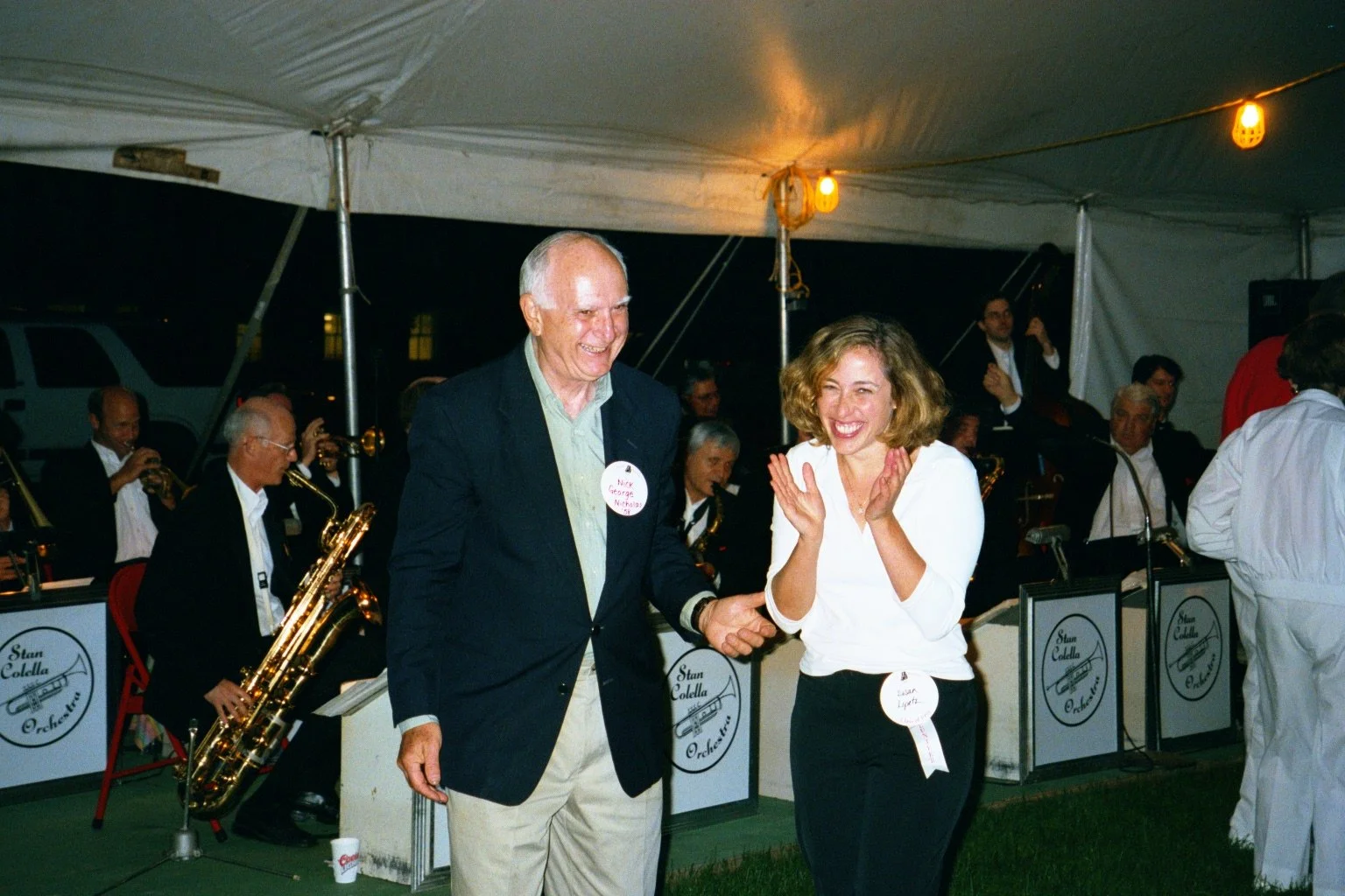 A woman and an older man smiling and holding hands at a band event under a tent, with musicians playing saxophones in the background during Cornell Reunion weekend.