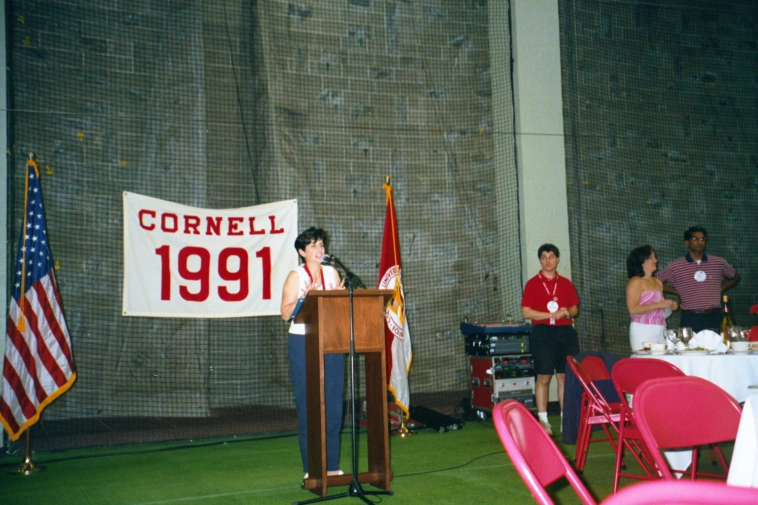 A woman is speaking at a podium during an event with a Cornell banner from 1991 in the background. There are three people standing nearby, and a round table with red chairs and table settings is in the foreground.