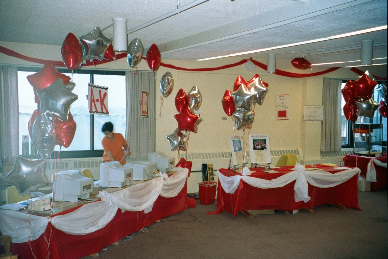 An indoor room decorated with red, white, and silver balloons, some shaped like stars, along with red and white tablecloths. There are tables with boxes, flyers, and a sign that says 'JOIN THE GROUP.' A woman in an orange shirt is working at a table,