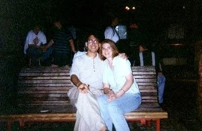 Two young women sitting on a bench at night, smiling, with people standing around in the background.