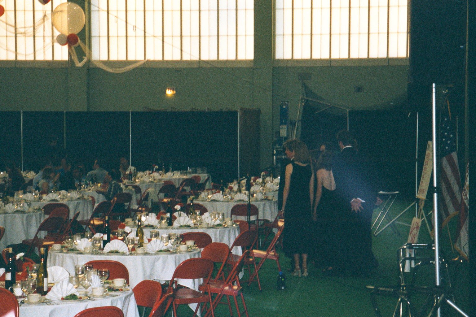 Indoor banquet hall with round tables set for a formal event, some guests seated, and a few people standing near a stage area with a backdrop and flags during Cornell Reunion weekend.