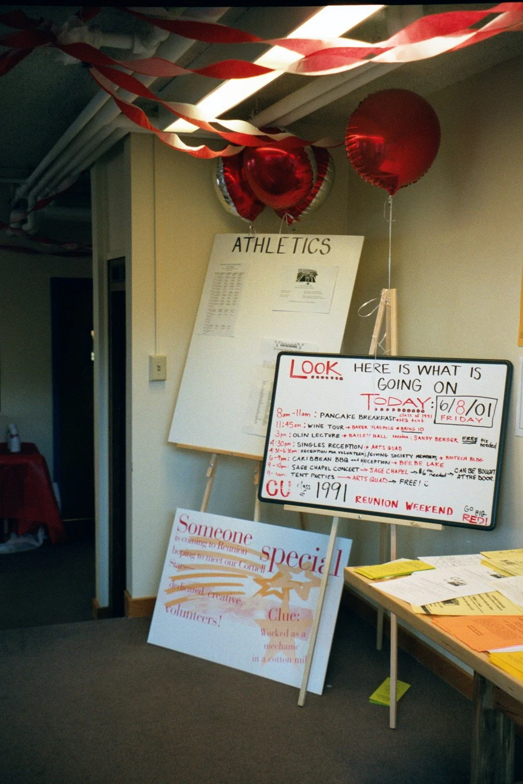 Decorated room with red balloons and crepe paper streamers, a white sign with 'ATHLETICS' on it, and a whiteboard listing events and schedule for a community event.