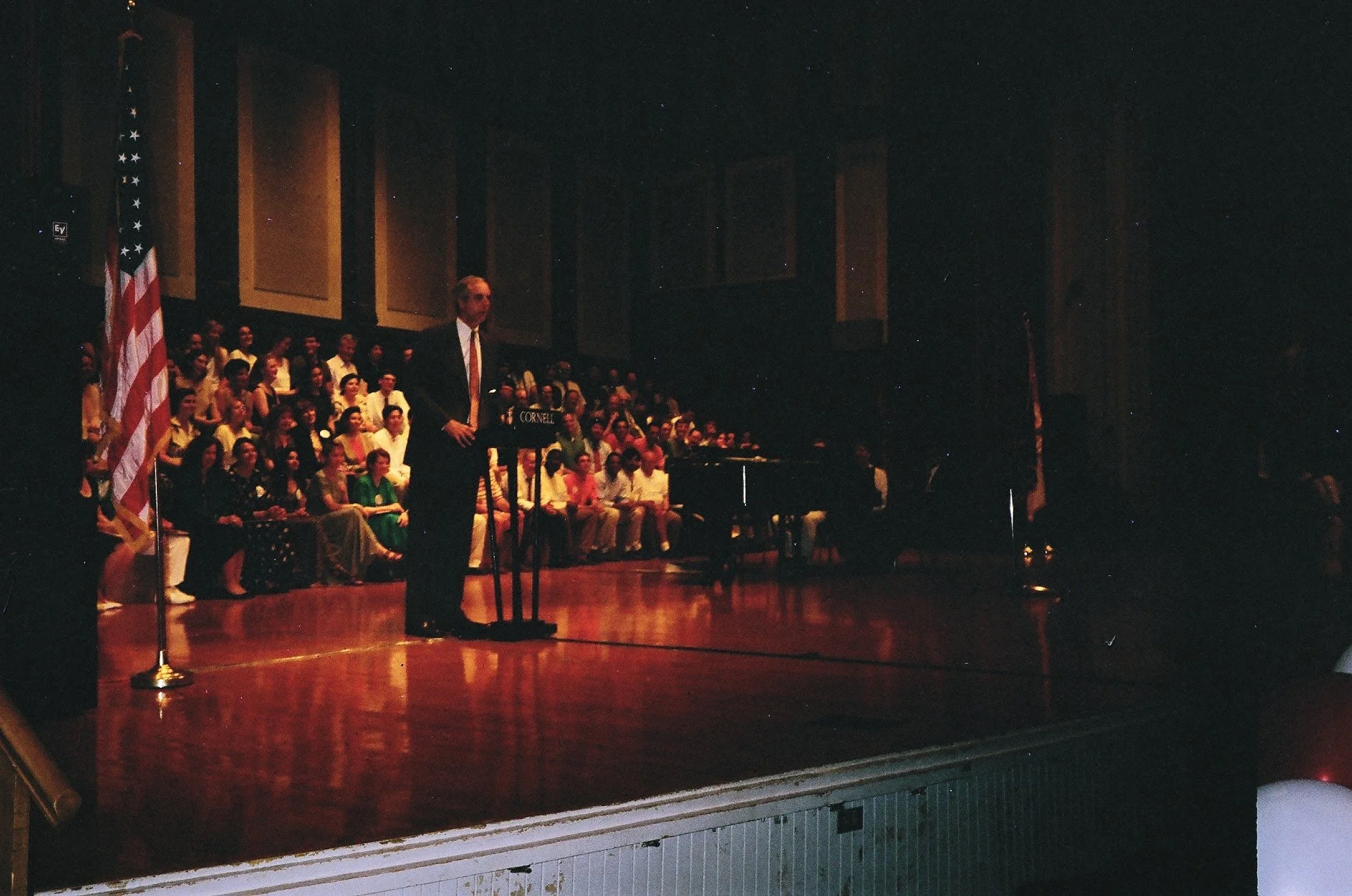 A man in a dark suit and tie standing at a podium on a stage with a large group of seated people behind him. There are American flags on either side of the stage.