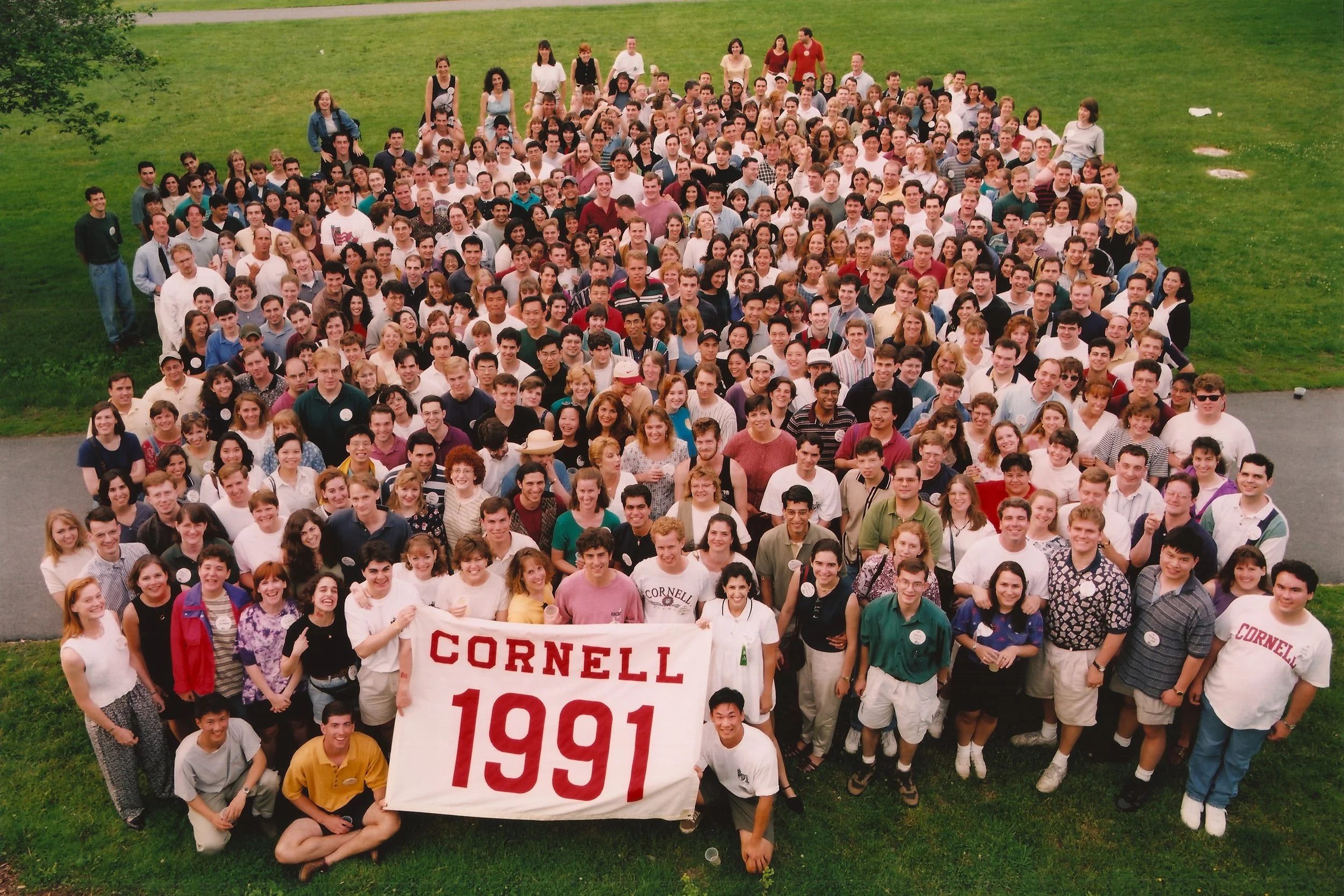 A large group of people gathered outdoors on a grassy field, holding a banner that says 'Cornell 1991,' suggesting a class reunion or similar event.