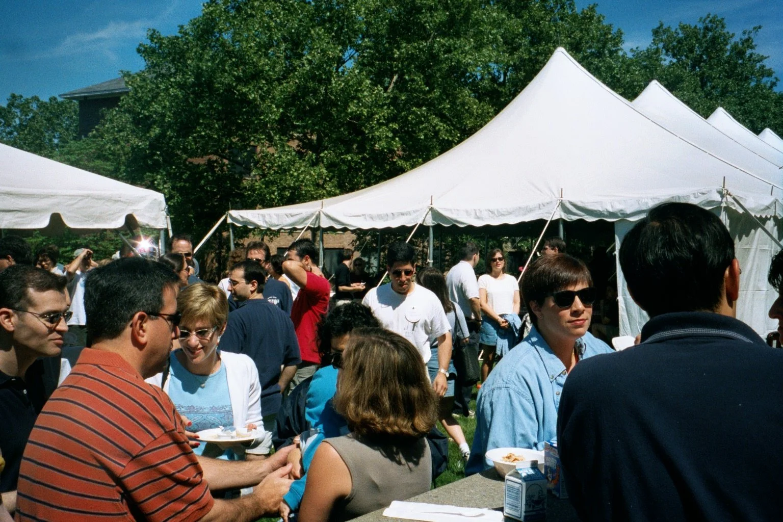 People gathered outdoors at a social event with white tents, green trees, and a blue sky.