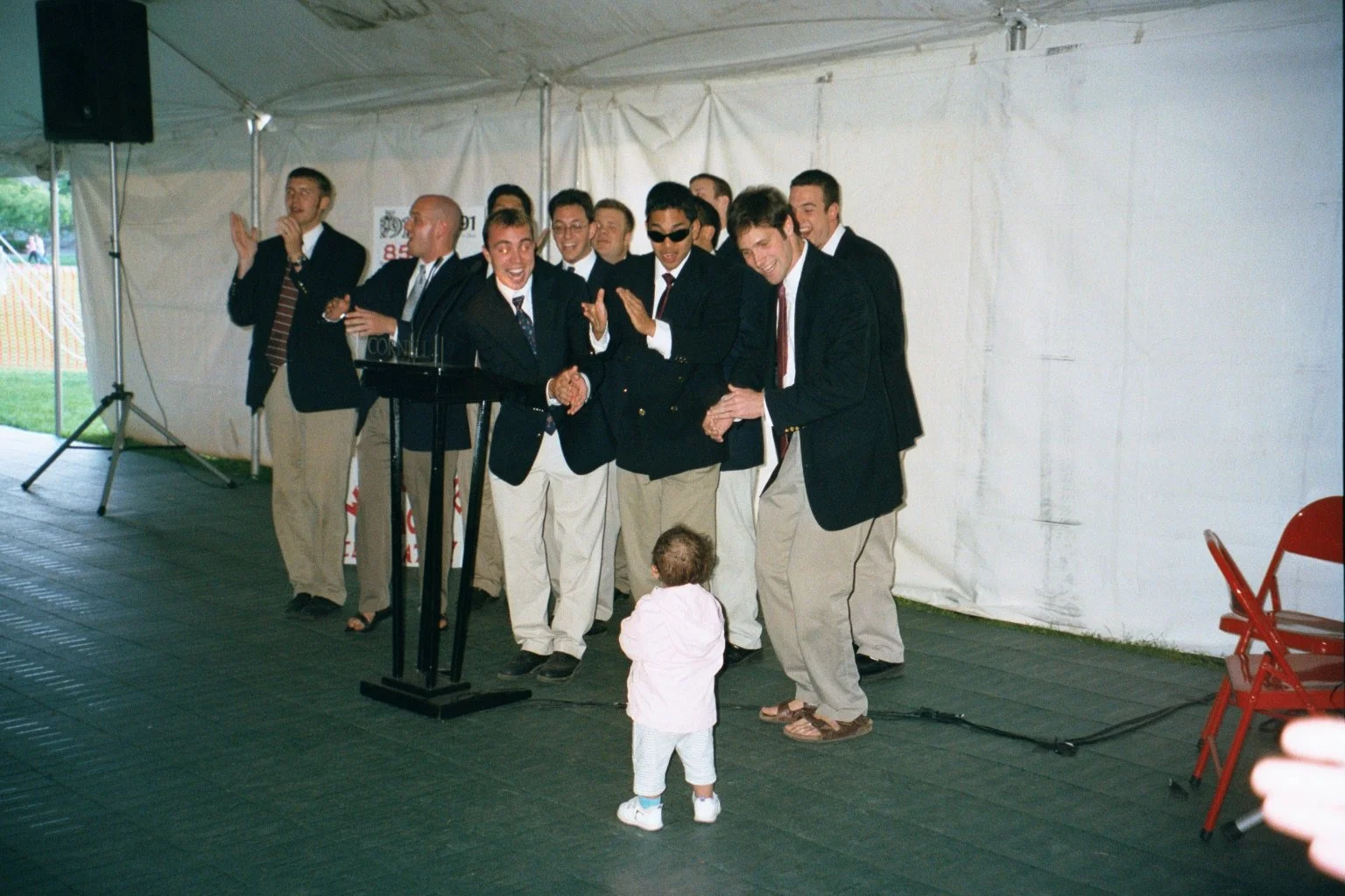 A group of men in suits, the Cornell Hangovers, some with sunglasses, are gathered around a podium, smiling, singing and clapping, under a white tent during Cornell Class of 1991 Reunion. A young child is standing in front of them, facing the group.