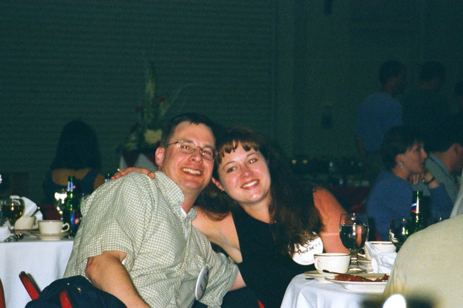 Two people smiling and leaning close together at a dinner party, with other guests seated at tables in the background.