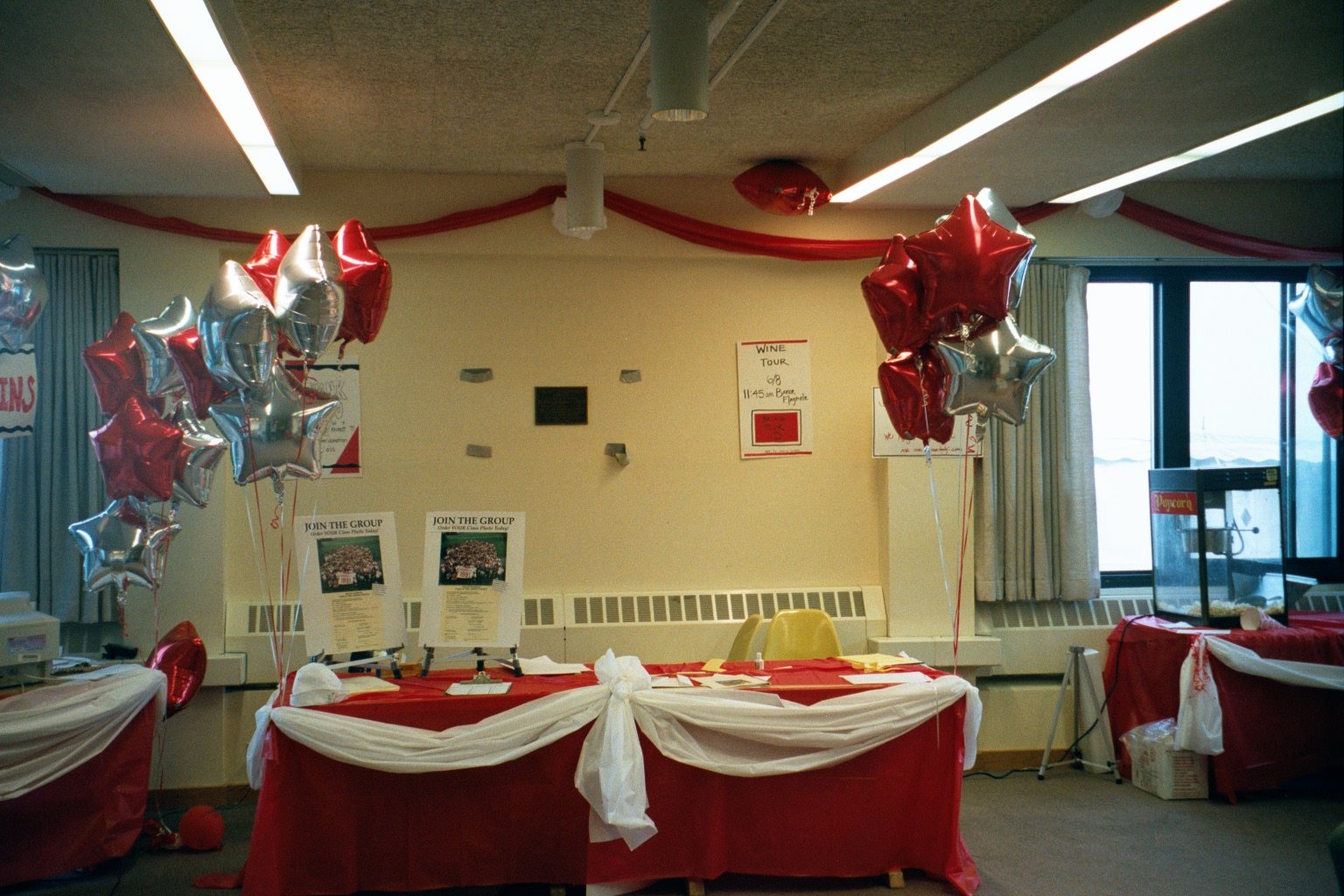 A decorated event space with red and silver star-shaped balloons, red and white tablecloths, and posters on the wall promoting a wine tour and group joining.