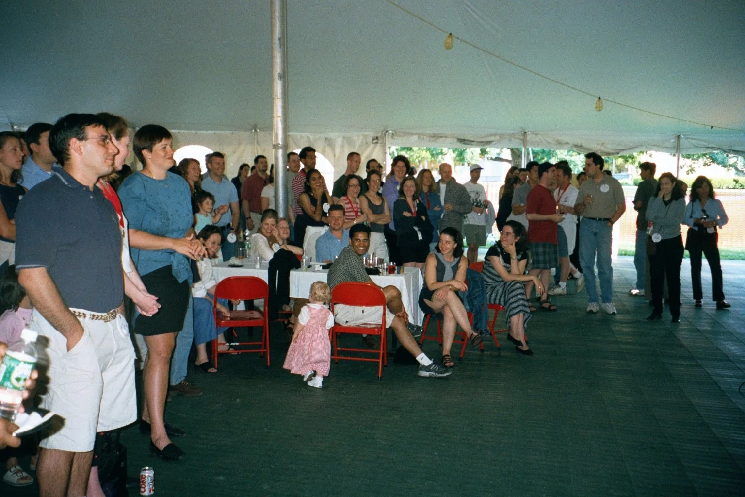 A group of people attending an indoor event under a large tent, some sitting at tables, others standing and socializing, with a diverse crowd including children.