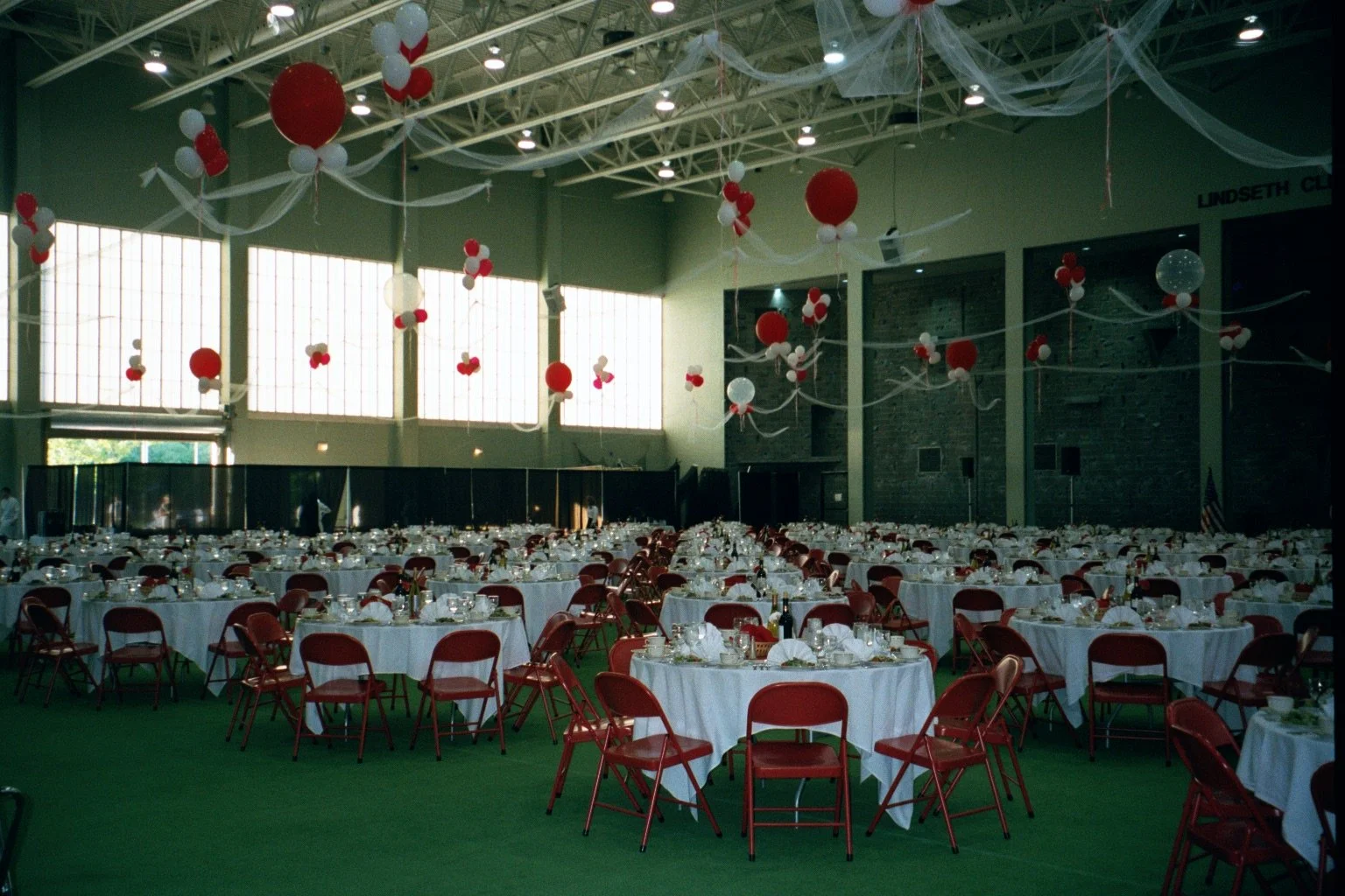 Large event hall decorated with white and red balloon centerpieces and streamers hanging from the ceiling, set up for a banquet with round tables covered in white tablecloths and red chairs, and a stage or back area in the background.