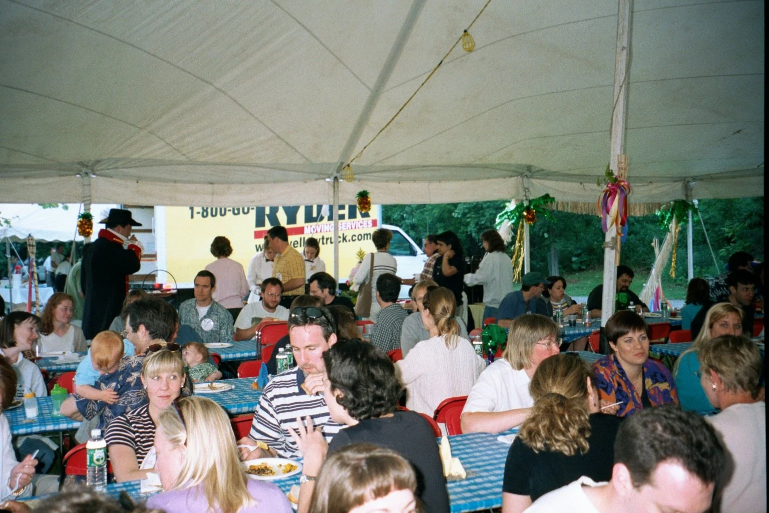 A large gathering of people seated at tables under a white tent at an outdoor event, with some standing and serving food, decorated with ribbons and ornaments.