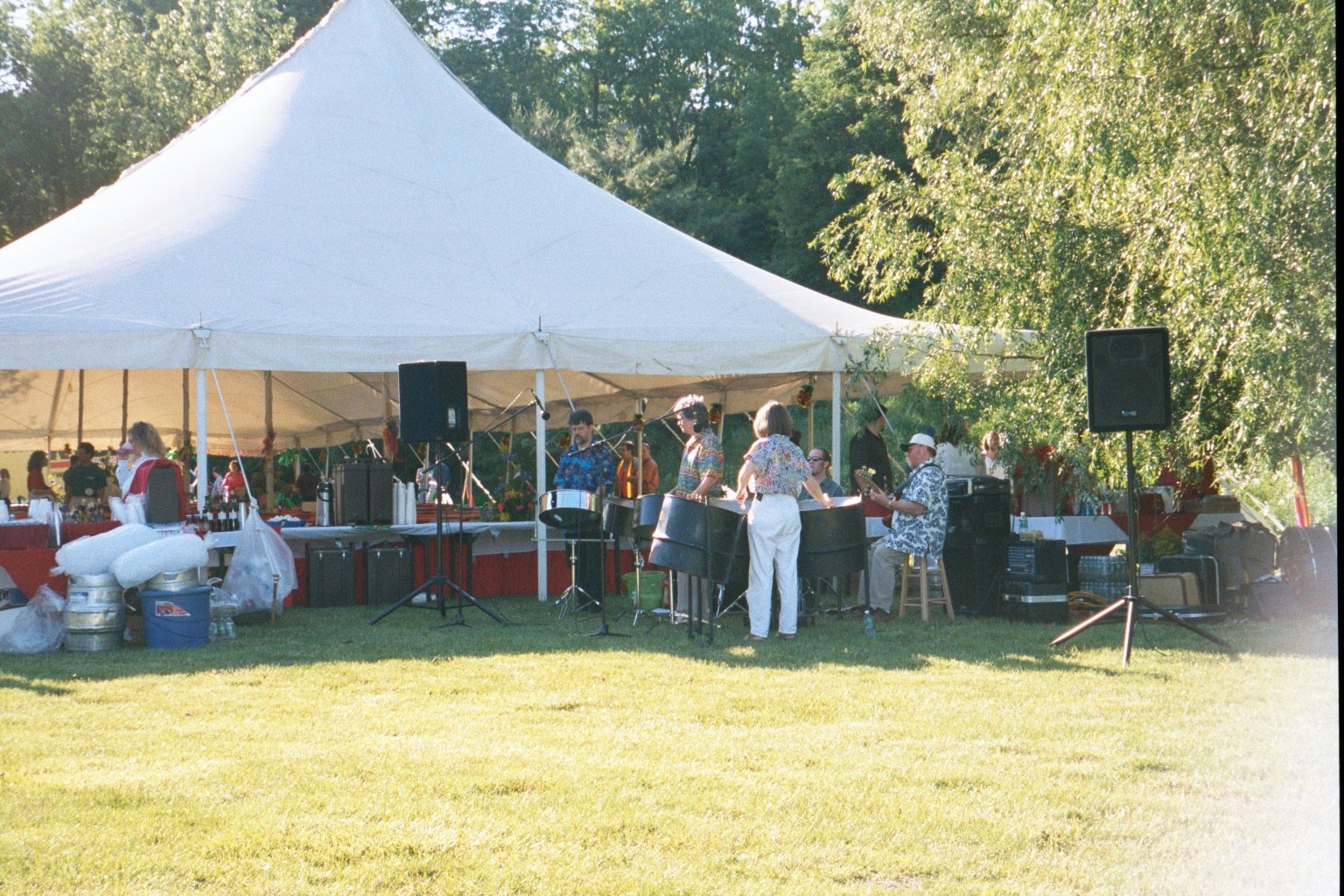 People playing steel drums under a large white event tent in an outdoor setting with green trees and grass.