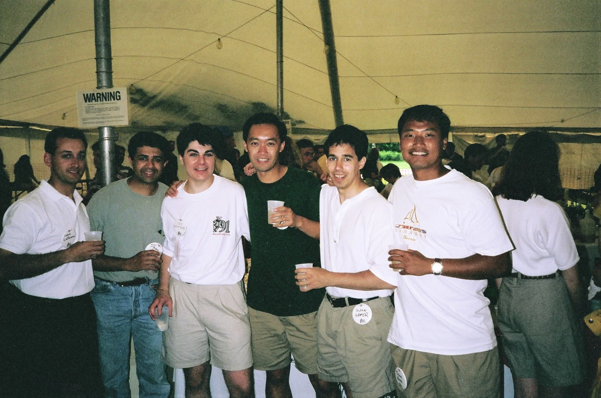 A group of six young men standing inside a large tent, smiling and holding cups to drink from, at an outdoor event.