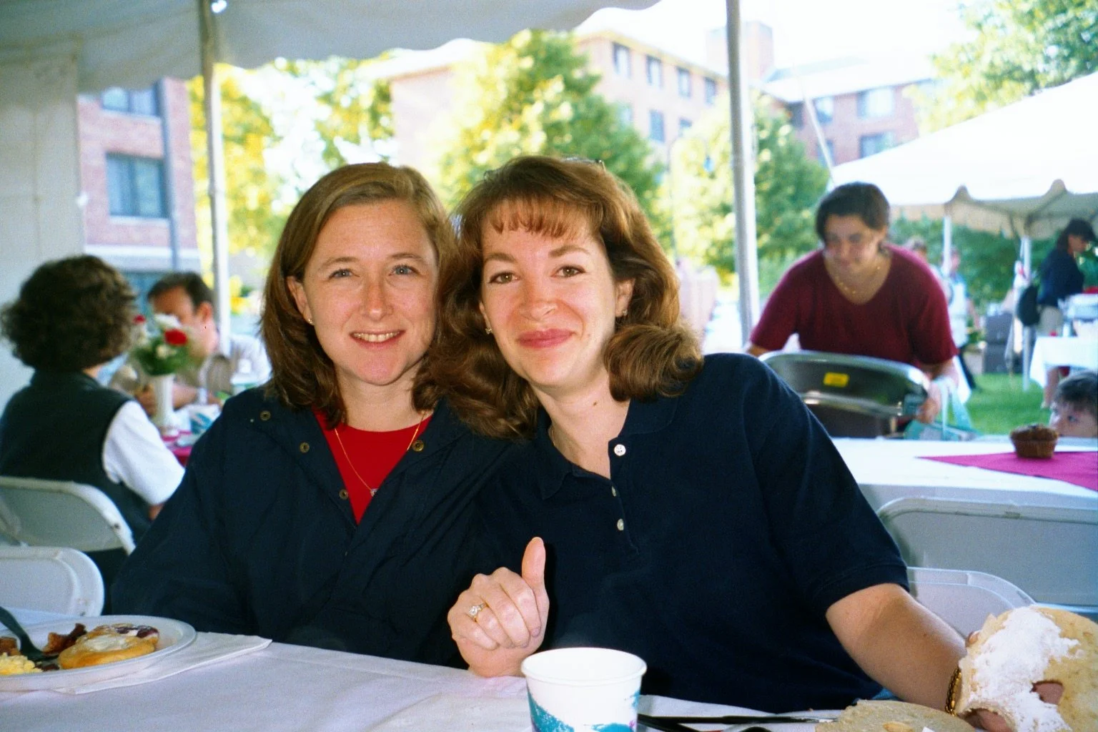 Two women sitting at a table, smiling, during an outdoor event, with other people and food visible in the background.