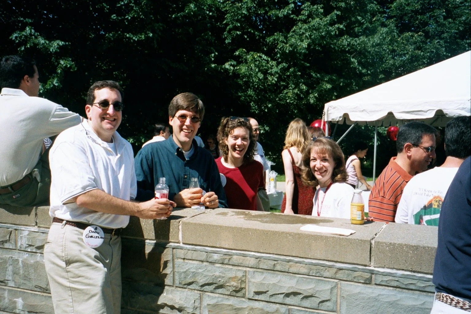 Group of people enjoying an outdoor gathering near a stone wall, with some holding drinks and smiling, under a tent with trees in the background.