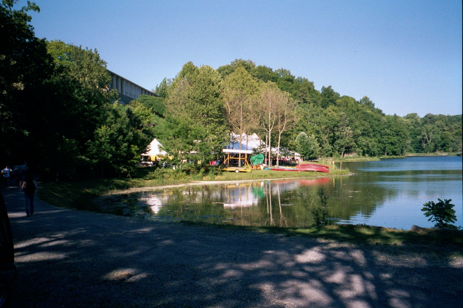 A lakeside scene at Beebe Lake during Cornell Reunion weekend with trees, tents, and boats on the shore, with reflections in the water.