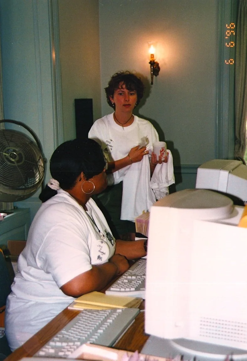 Two women working at a computer station. One woman is sitting and typing on a keyboard, wearing glasses and earrings, while the other woman stands nearby holding a plastic cup and a paper towel. There is a vintage computer monitor and keyboard, a fan
