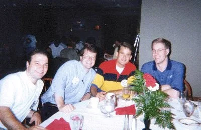 Four young men sitting at a banquet table with white tablecloths, red napkins, and a floral centerpiece, during a social event or dinner.