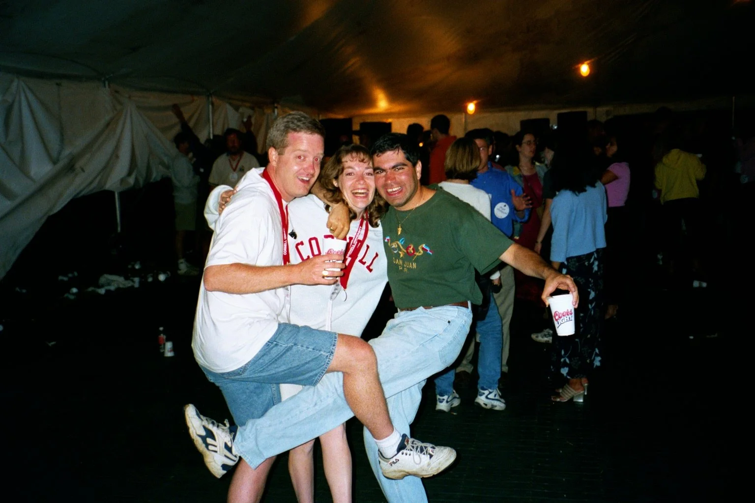 Three friends at a party, during Cornell Reunion weekend, two men and one woman, smiling and celebrating, with drinks in their hands, surrounded by other party-goers.