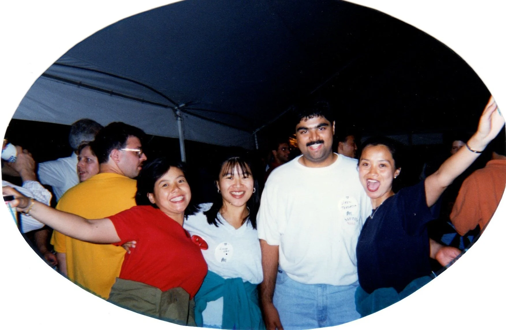 Group of five smiling people at a social event under a tent, with three women and one man in the foreground, one woman with arms outstretched, all appearing happy and celebrating.