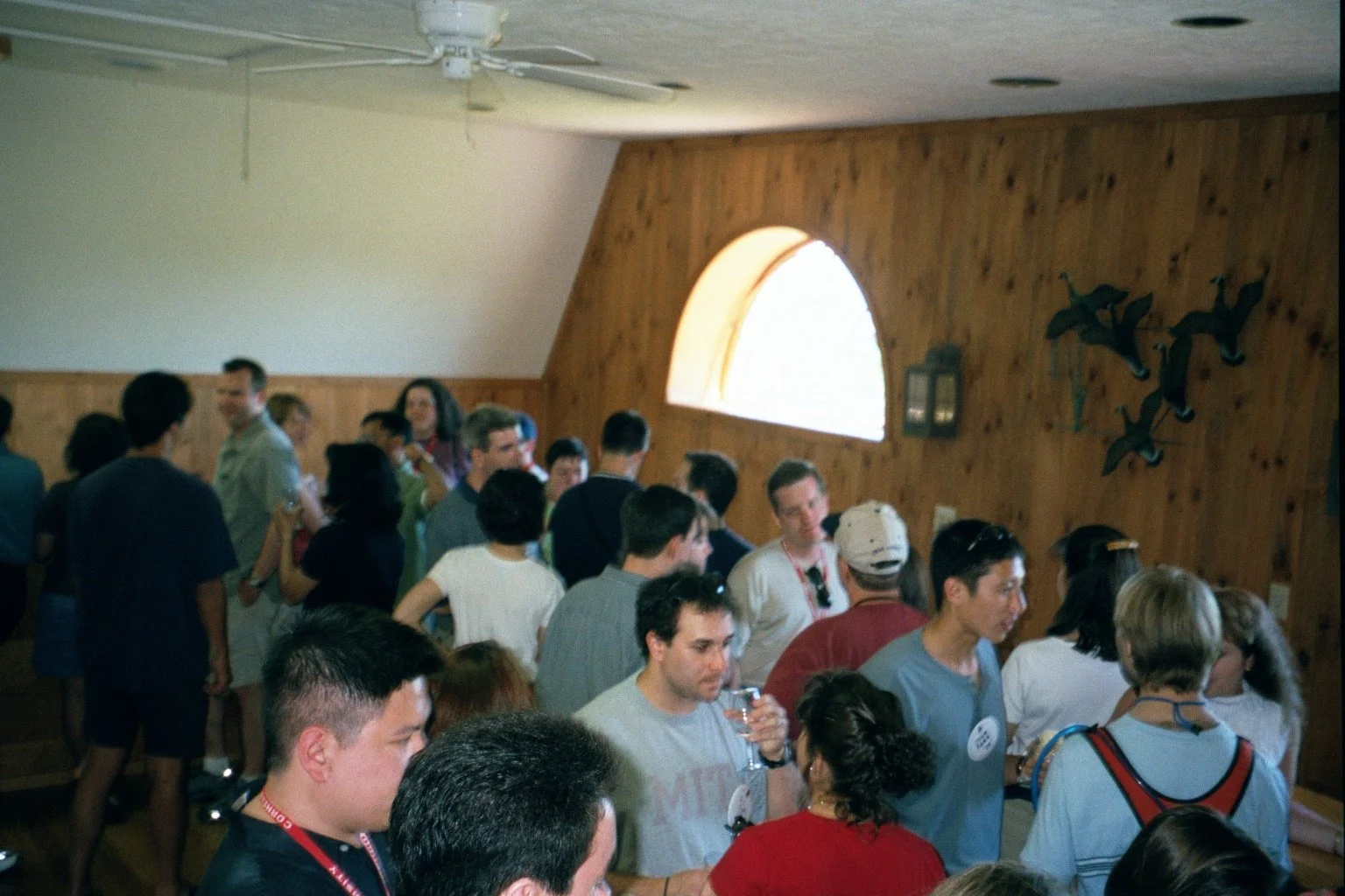 A crowded indoor gathering with people socializing, some holding drinks, in a room with wooden walls and a large arched window.