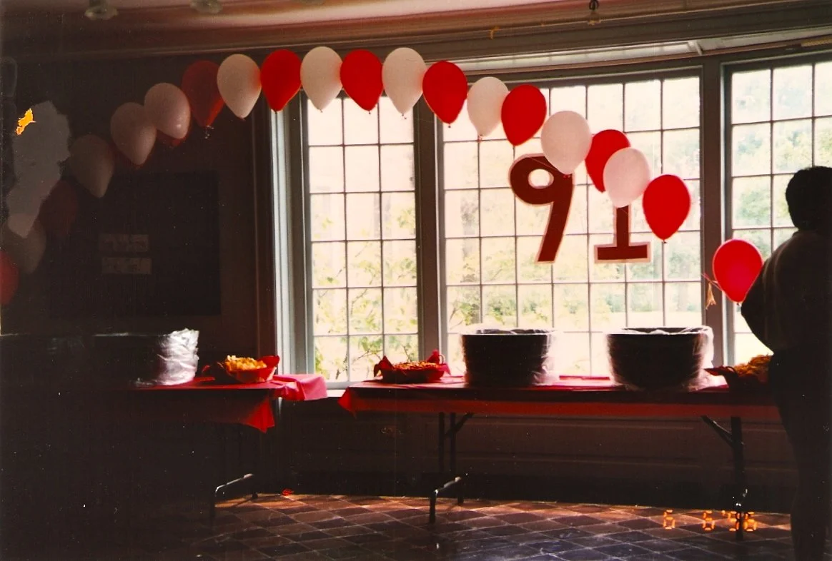 Decorated celebration table with balloons, large cutouts of the number 91, and bowls of snacks near a large window.