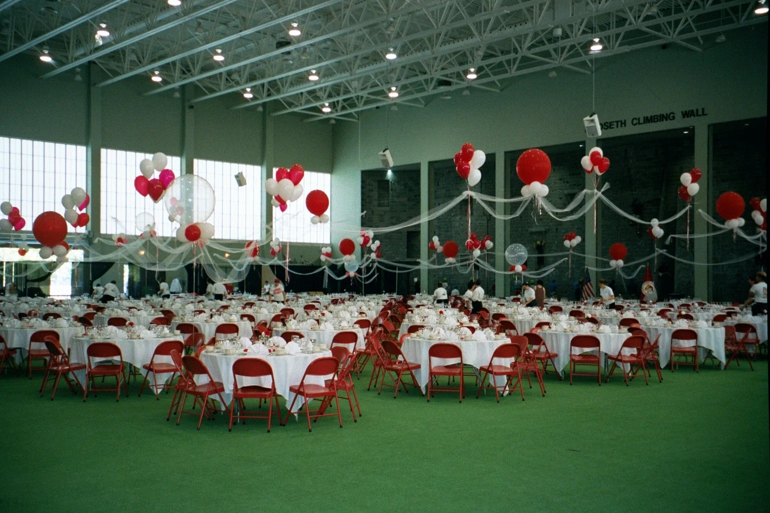 Large indoor event space decorated with red and white balloons, tablecloths, and chairs, set up for a celebration or banquet.
