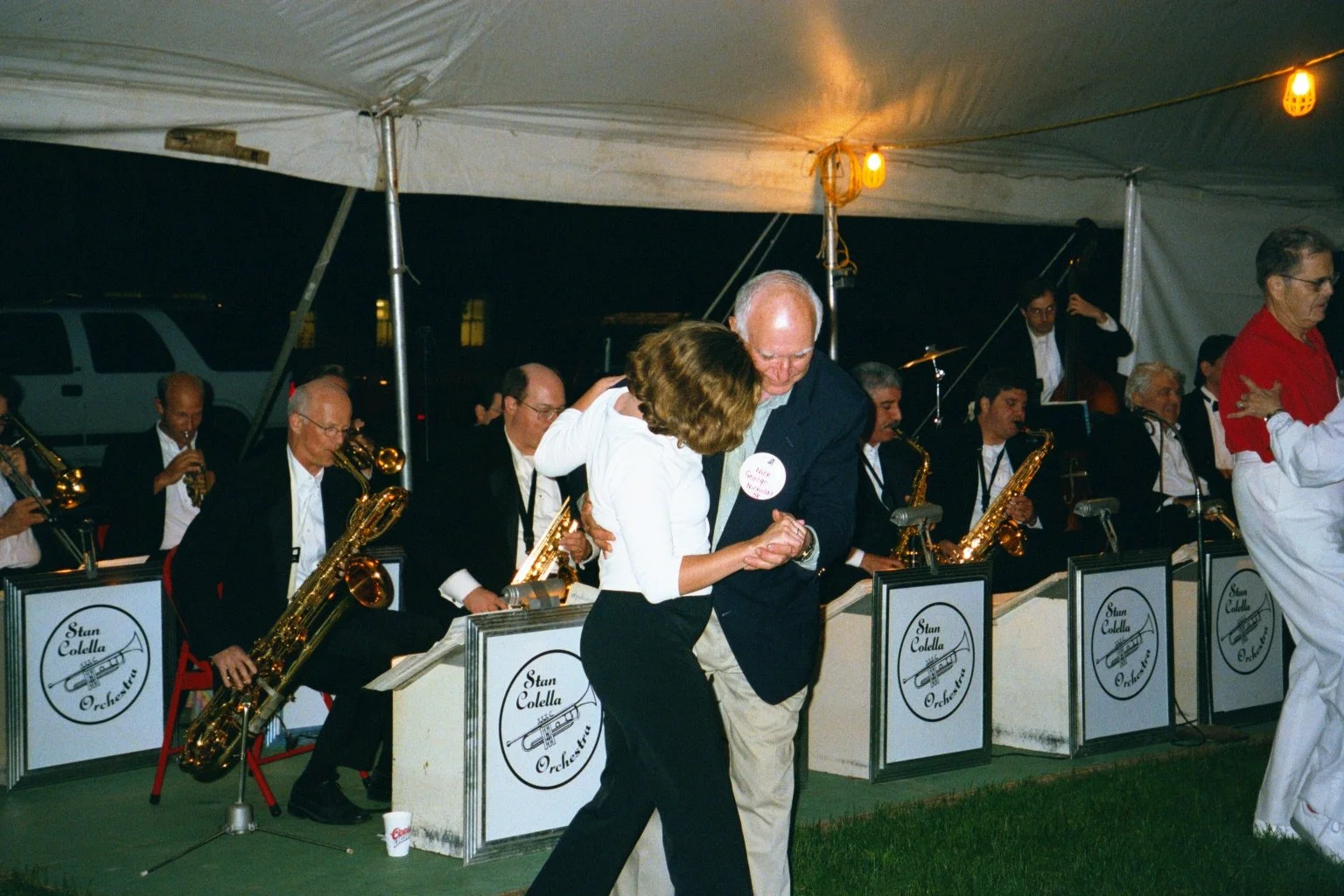 Two people dancing in front of an orchestra at an outdoor event. The orchestra members are sitting with saxophones, and the band sign reads 'Stan Colella Orchestra.' The scene is under a tent at night during Cornell Reunion weekend.