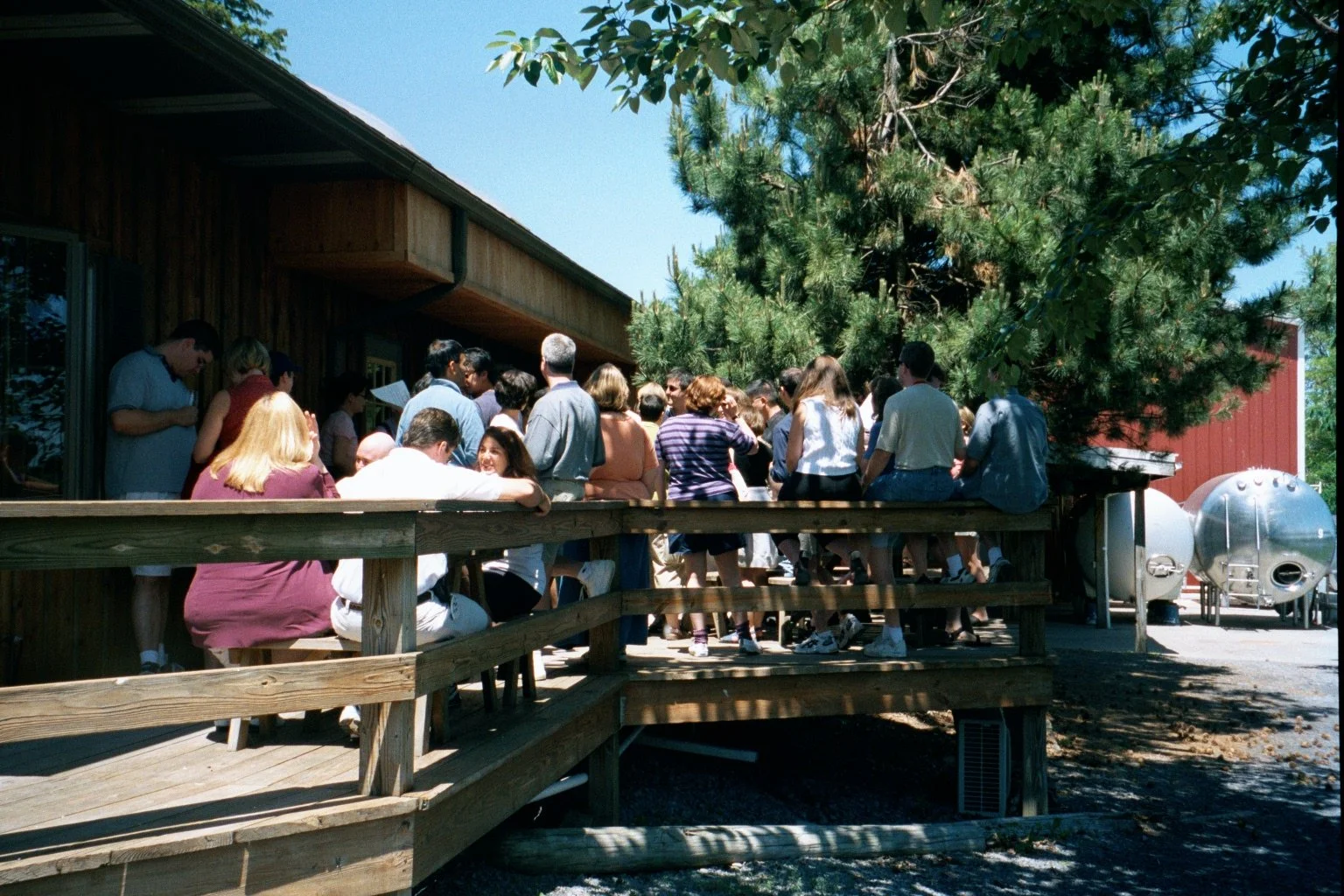A gathering of people on an outdoor wooden deck next to a wooden building, with some people standing and others sitting, and a large tree providing shade.