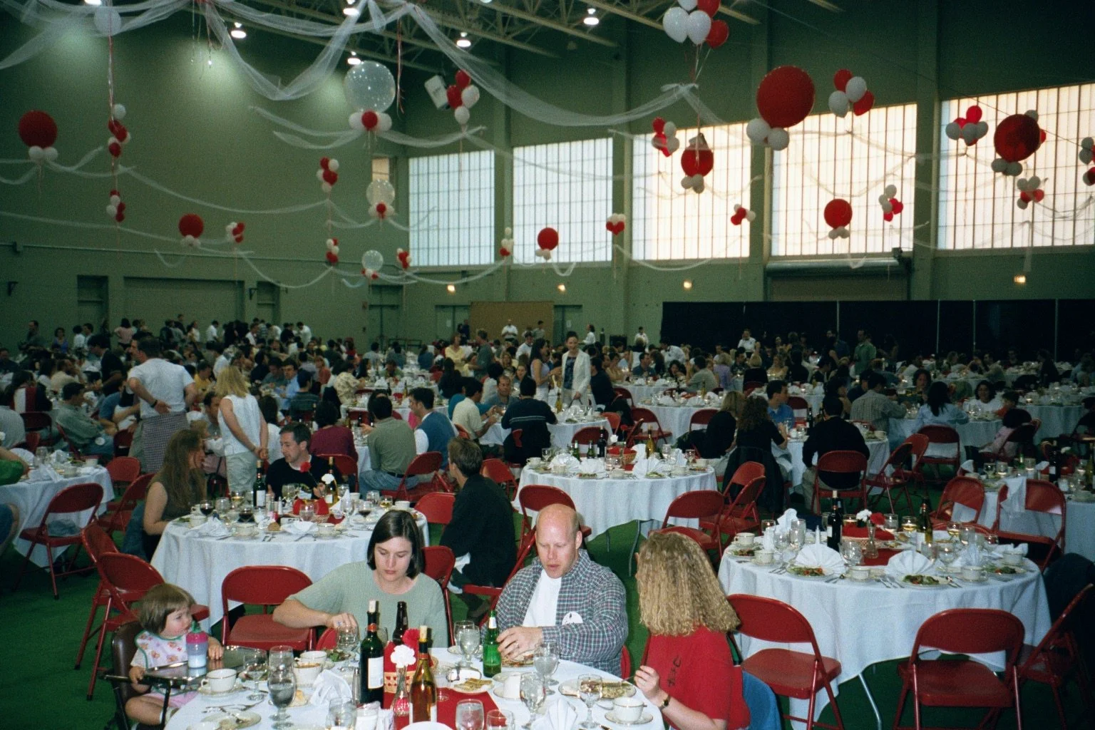 A large indoor banquet hall decorated with red and white balloons and streamers, filled with many round tables set for a meal, with guests dining and socializing.