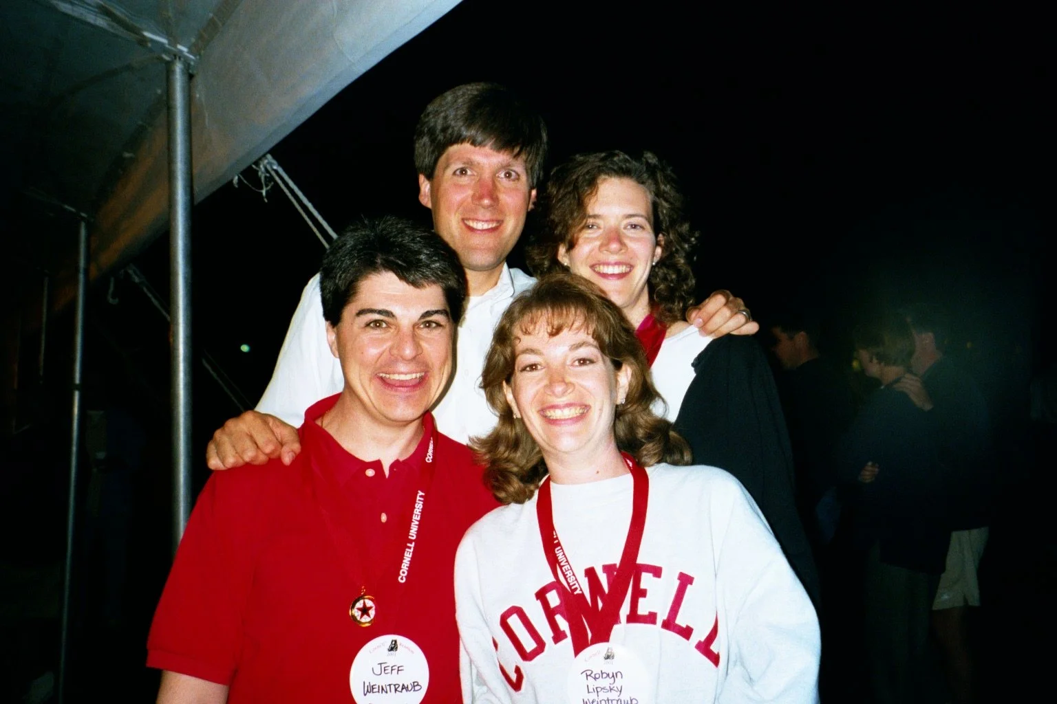 A group of five people smiling and posing together at an event, with some wearing Cornell University shirts and name tags during Cornell Reunion weekend.