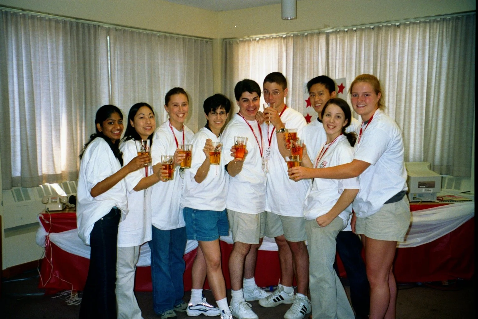 A group of eleven young people celebrating together indoors, holding glasses filled with a drink, smiling, and wearing casual clothes with some in white shirts and medals around their necks.