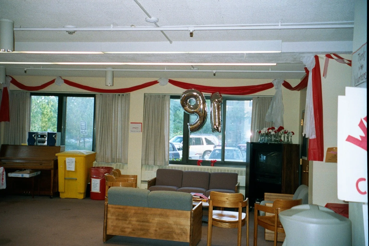 Celebration area with silver balloons forming the number 91, red and white streamers hanging from the ceiling, a brown sofa, wooden chairs, and a small black cabinet with flower vases on top. Large windows with curtains show parked cars outside.