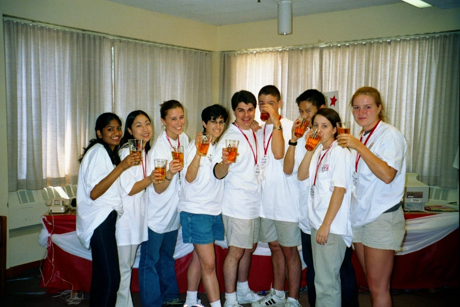 A group of nine young adults, dressed in white medical uniforms, standing together in a room with beige curtains, holding glasses of amber-colored drink and smiling for the camera.