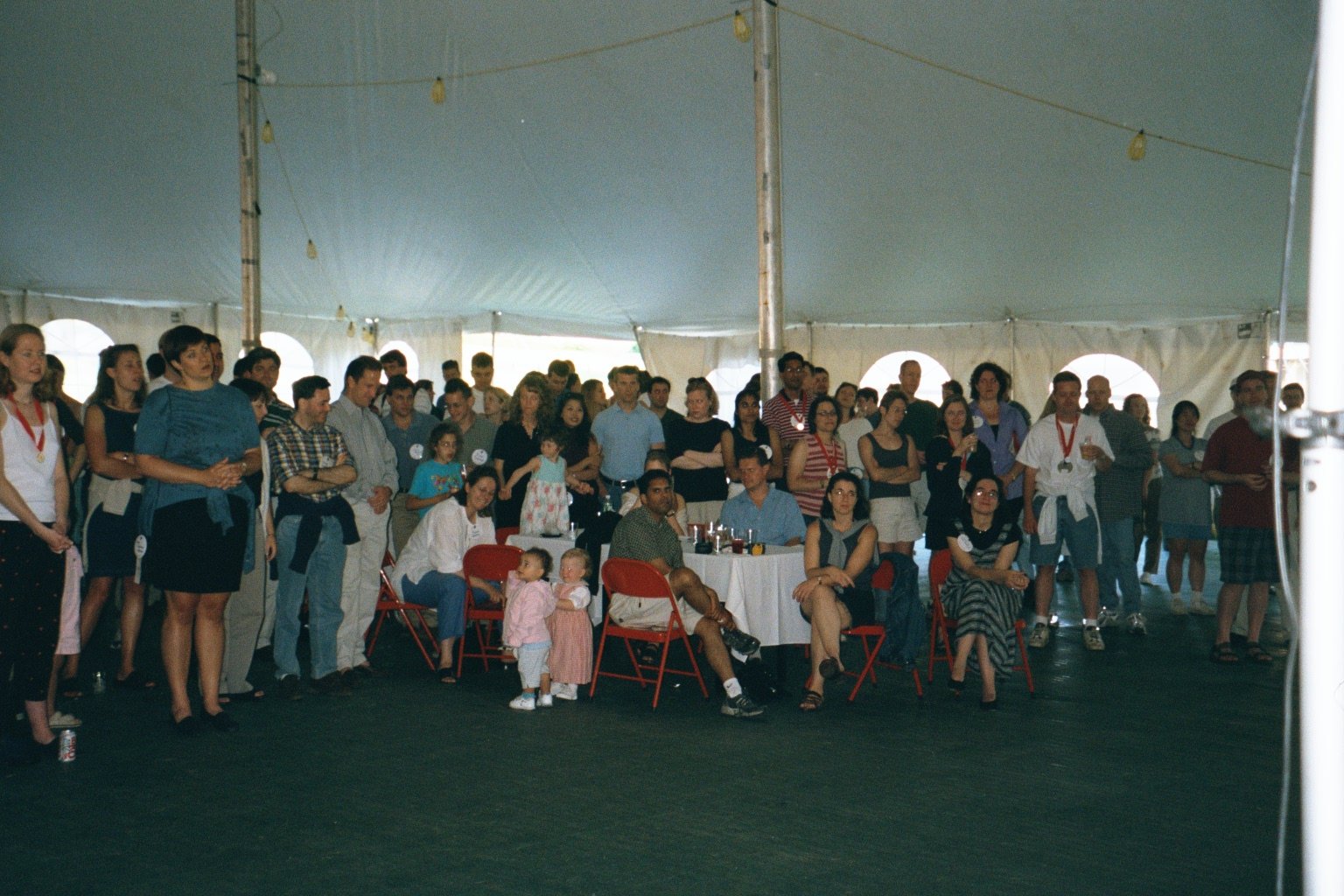 Group of people inside a large white event tent, some standing and some sitting, attending a social gathering or party.