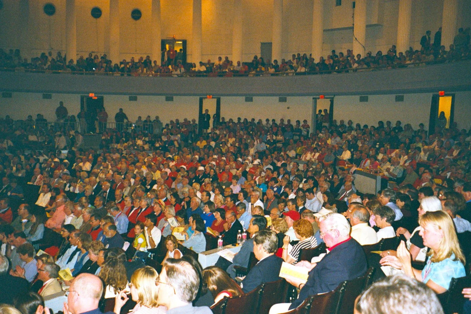 A large audience seated in Bailey hall, attending Cornelliana Night during Cornell Reunion weekend. The seating fills the auditorium, with some people on the balcony level. Many are engaged, reading, or using phones.