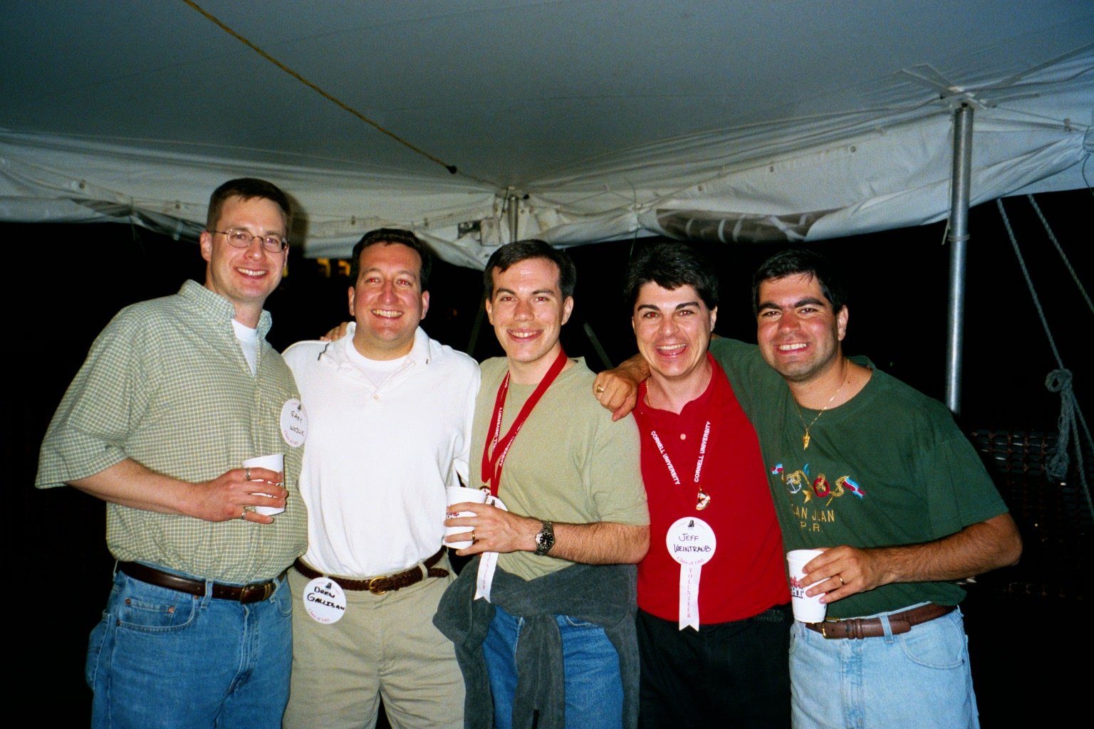 Five men standing together at a nighttime event, smiling, with medals and cups, under a tent during the tent parties at Cornell Reunion weekend.